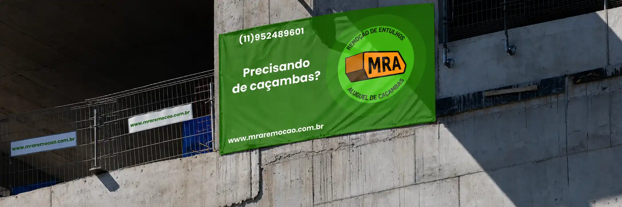 Parede de prédio em construção com a cor cinza, onde há quatro banners, um deles em destaque por ser o maior, e de uma cor diferente: ele é retangular verde escuro, contendo em branco um número de telefone, o título 'Precisando de caçambas?' e o URL 'www.mraremocao.com.br', nesse mesmo retângulo também se encontra a logo do MRA remoção, envolta em dois círculos verdes claro. Sobre a logo, se trata de uma caçamba amarela com as iniciais 'MRA' em sua lateral, talbém em forma de cículo se encontram duas frases em verde escuro: 'remoção de entulhos' e 'aluguel de caçambas'. Sobre os três banners menores, eles são de cor branca e todos contém a URL do site 'www.mraremocao.com.br'
